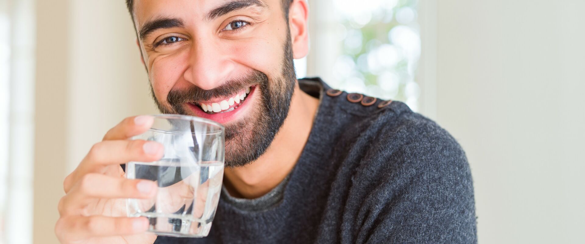 Ein Mann mit leichtem Bart lächelt freundlich und hält ein Glas Trinkwasser in der Hand.