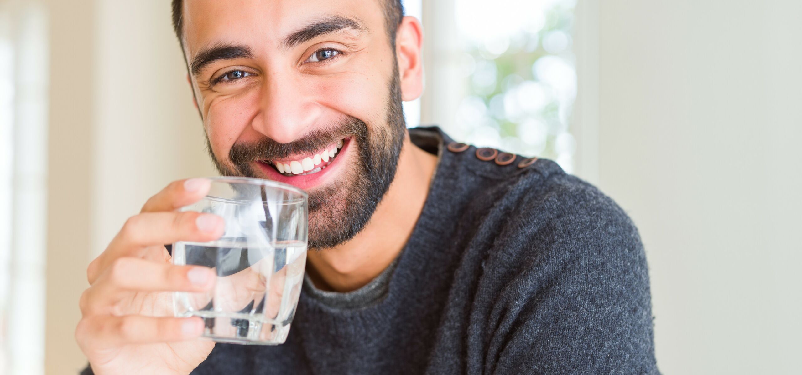 Ein Mann mit leichtem Bart lächelt freundlich und hält ein Glas Trinkwasser in der Hand.