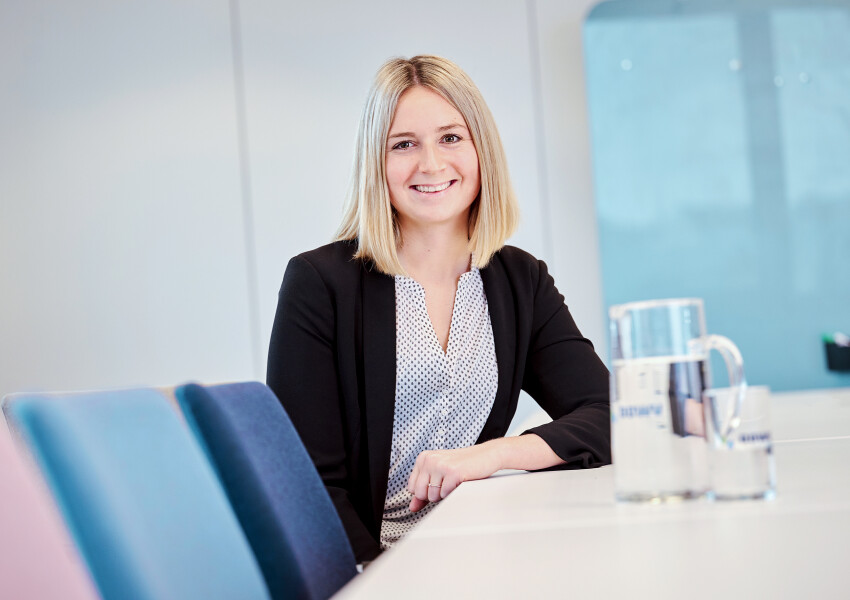 Recruiterin Michaela sitzt im Konferenzraum am Tisch und lächelt freundlich. Vor ihr steht ein Glaskrug mit frischem Trinkwasser.